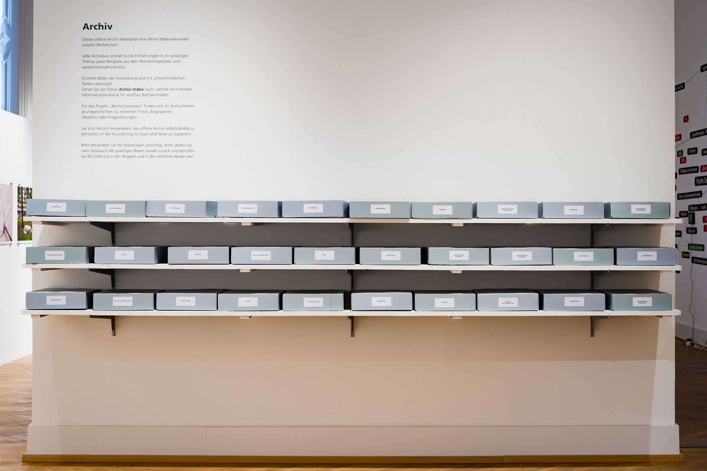 Archive boxes organized on shelves in a white exhibition space. Above them on the wall a text in German with the title Archiv (archive).