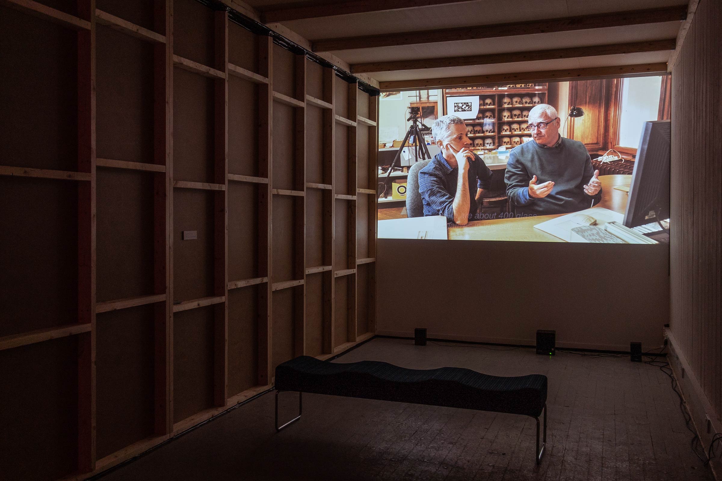 A dark exhibition space with a large video projection on the wall and a bench in front of it.