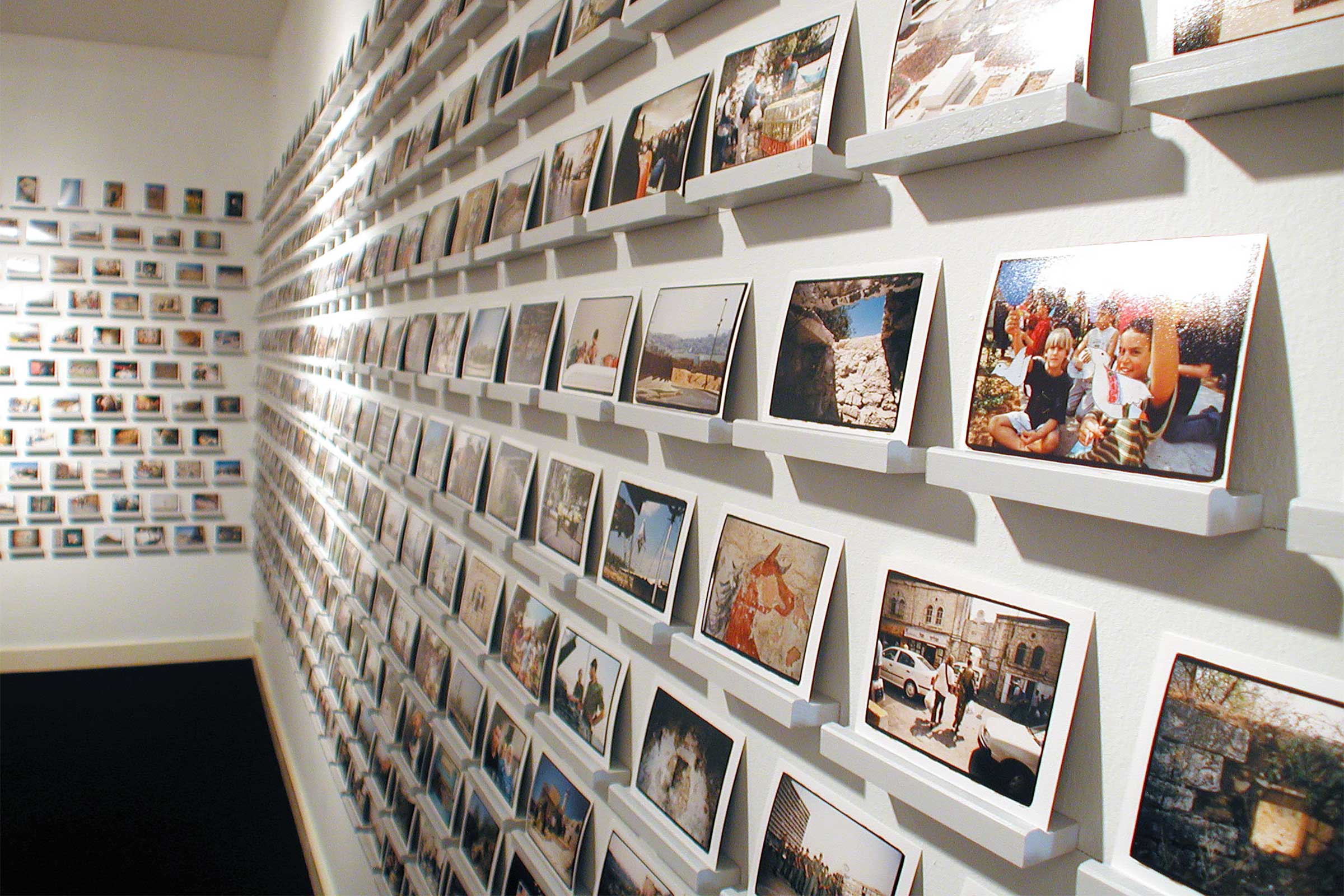 White exhibition space covered with hundreds of small photographic prints on individual small shelves directly mounted on the wall.