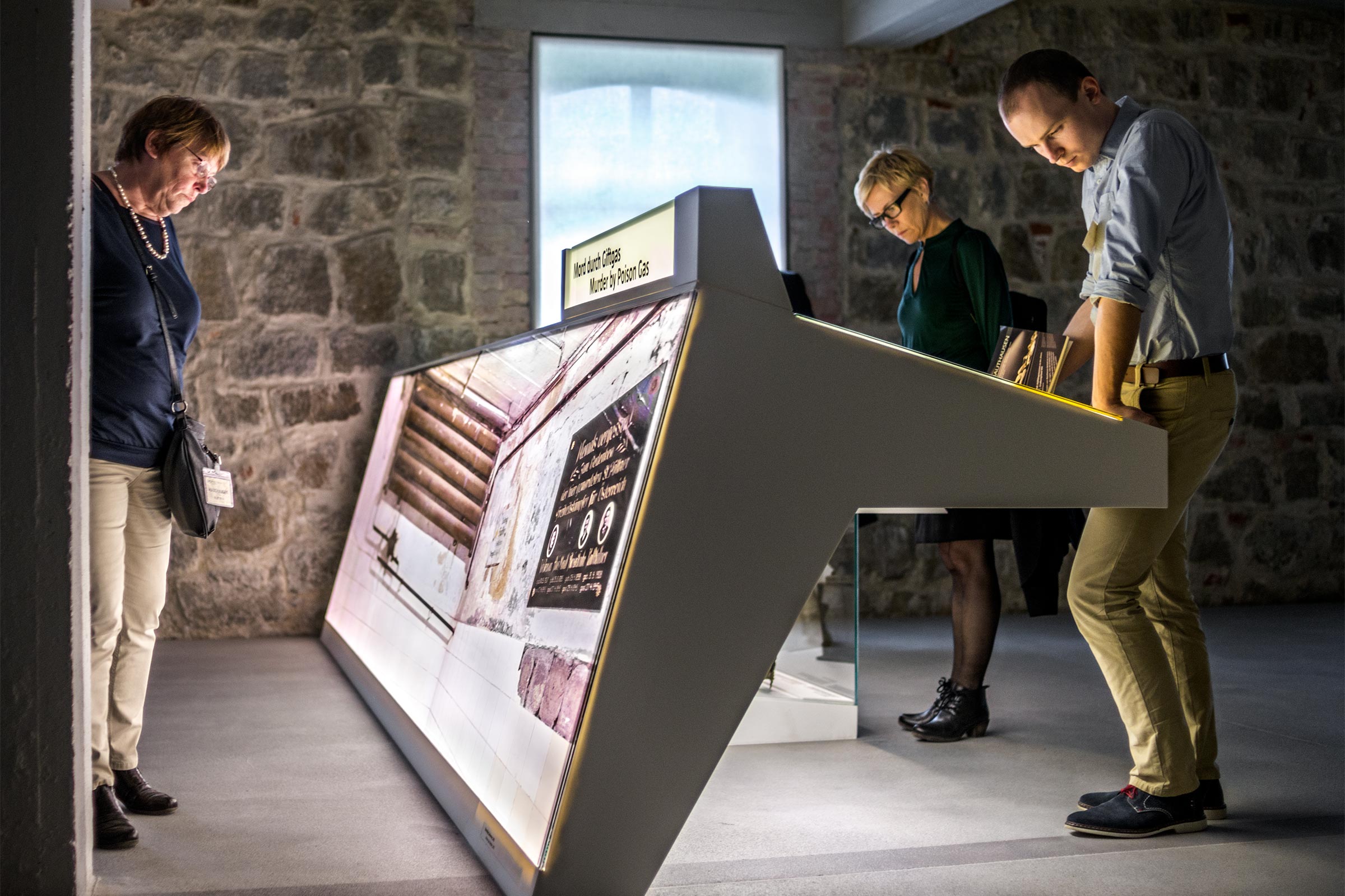 Three people looking at an exhibition display consisting of a large back-lit photograph on one side and a lit table on the other. The label reads Murder by Poison Gas in English and in German.