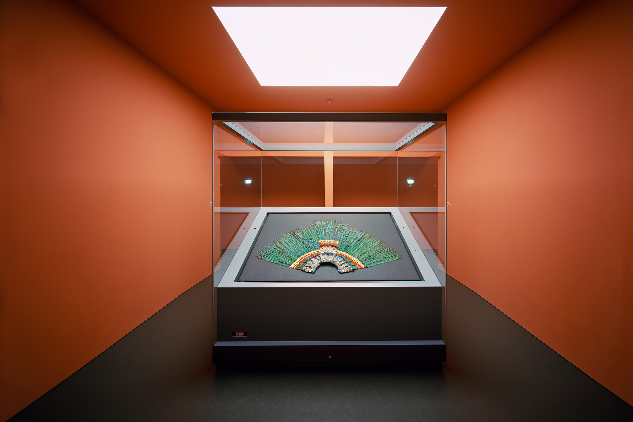 Large display case in a museal space painted organge. Inside the case is a large feathered head-dress with golden decorations and long green feathers.