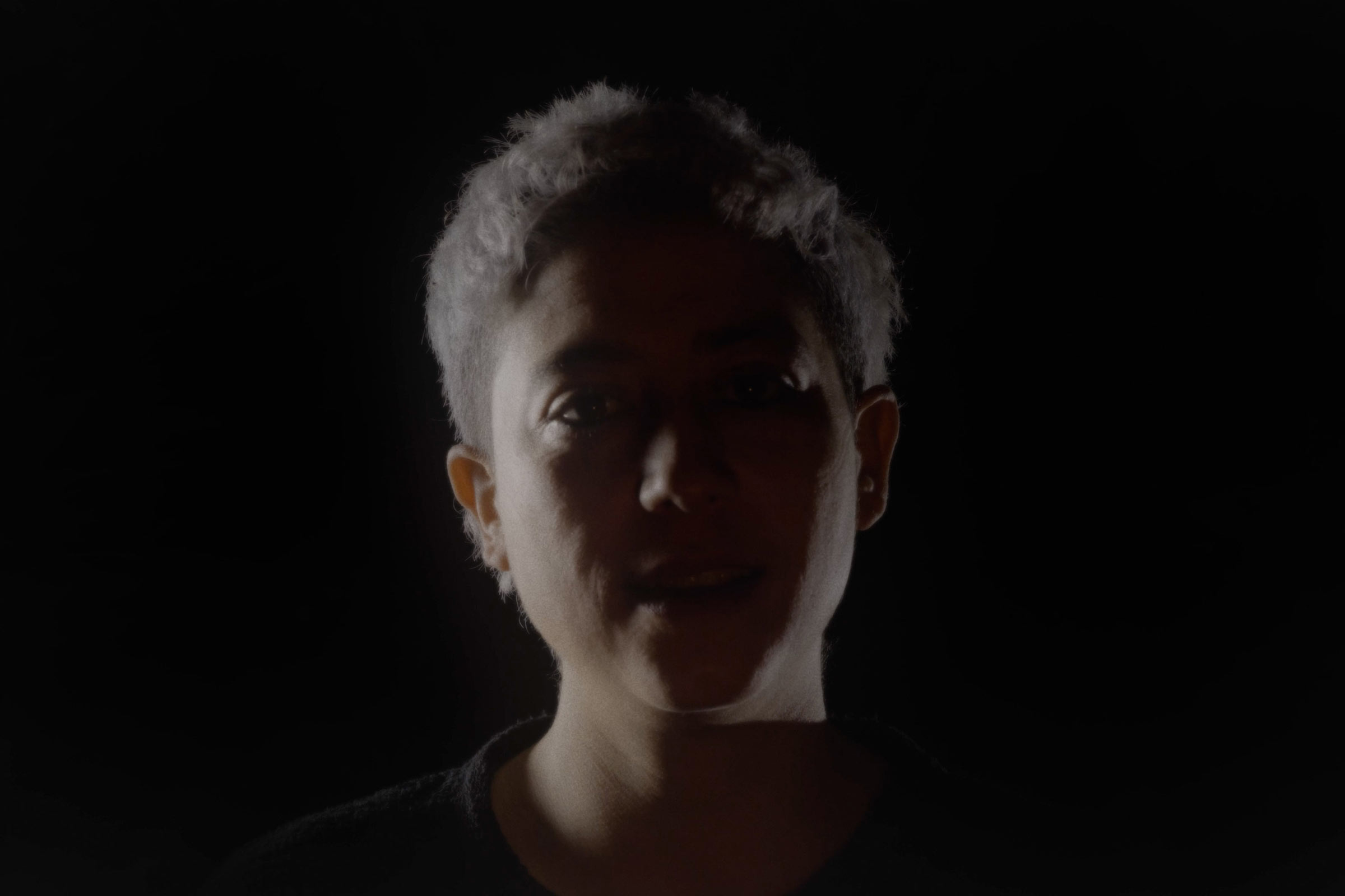 Woman's face, mostly in the shadow, dimly lit from the sides, against a dark black background.