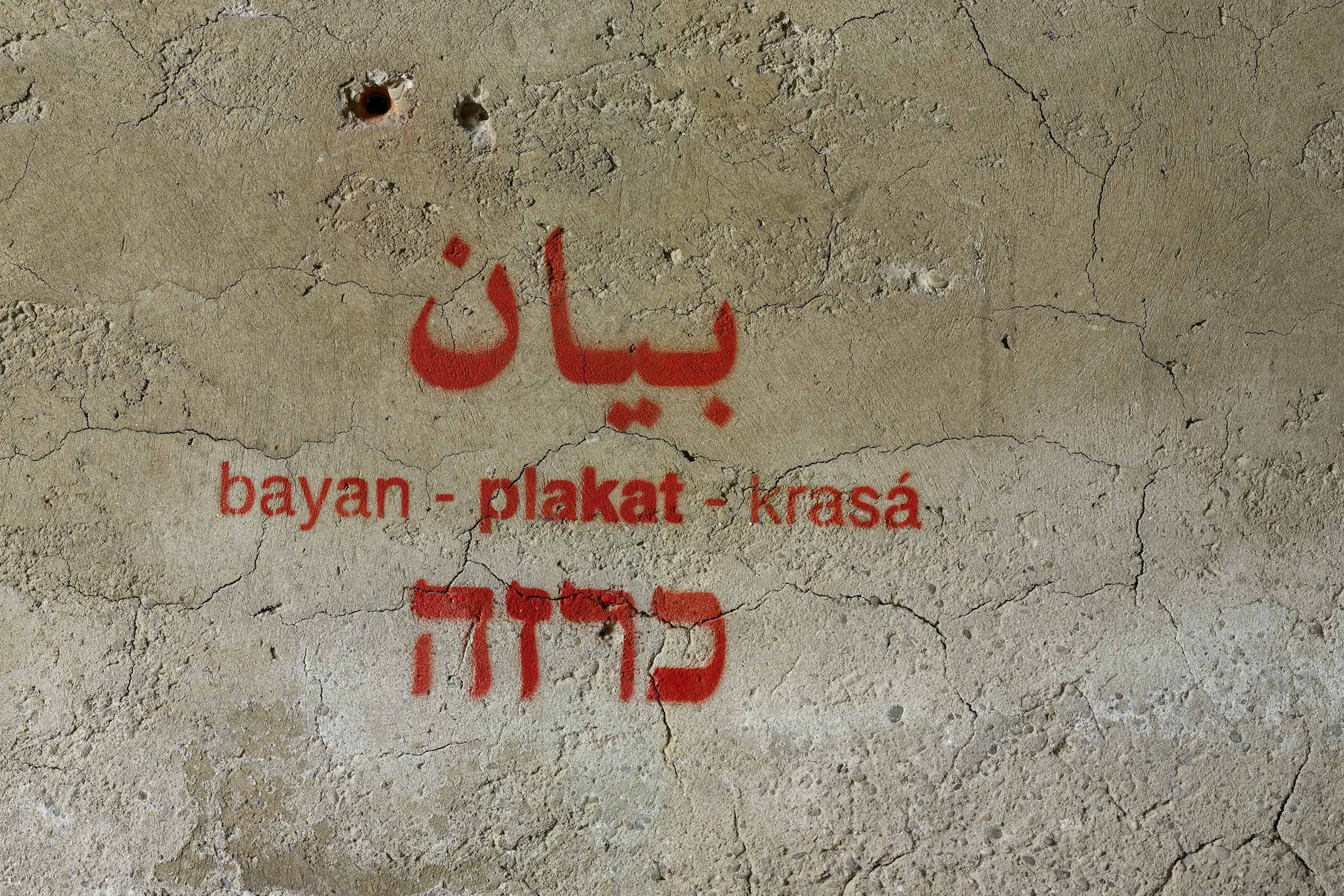 a fragment of rough concrete wall with stencil-graffiti text in red of the word poster in Hebrew, Arabic, and their transliteration in German: 'bayan - krasa', as well as the German word 'plakat'.