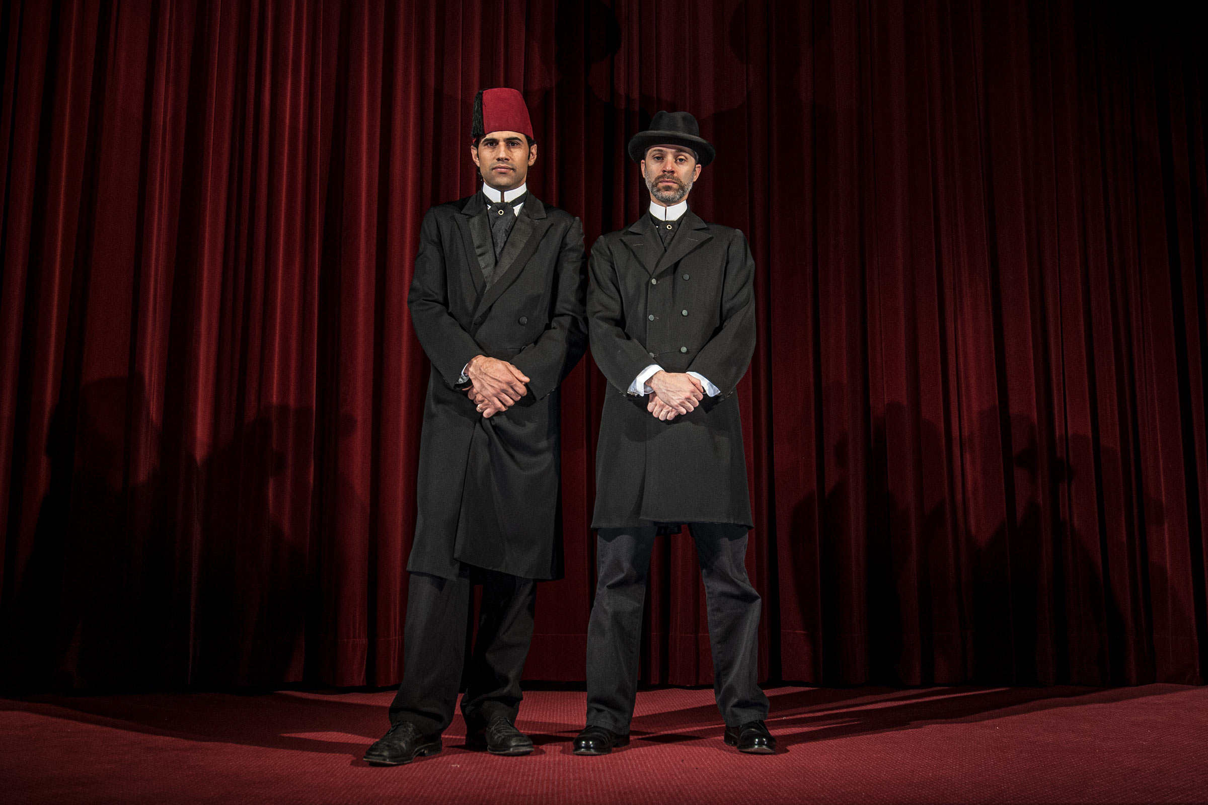 Two men standing on a stage with a red curtain behind them wearing black, 19th century suits. One of them is wearing a traditional red Fez hat and the other a European black dress hat.