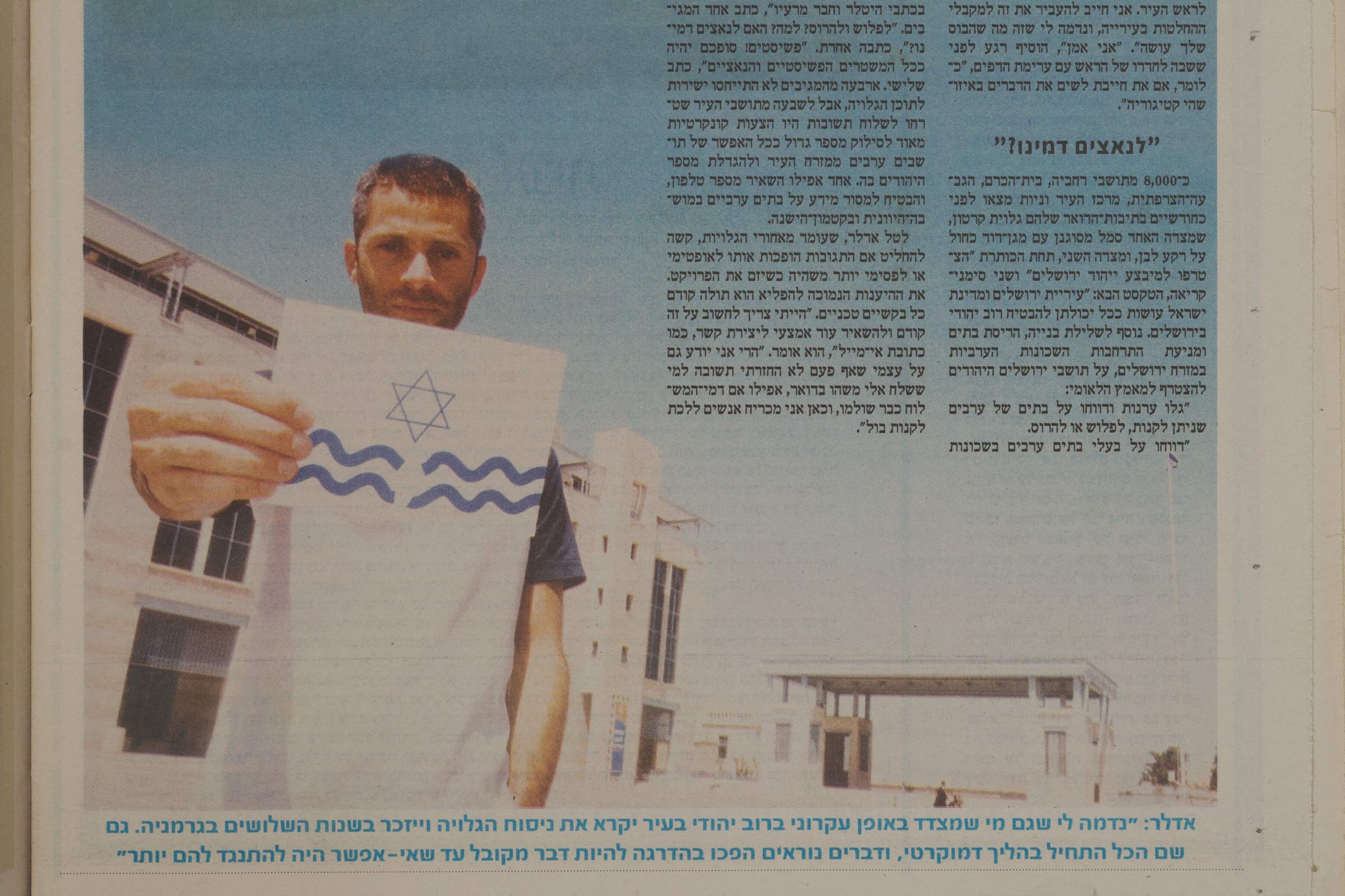 Newspaper clip with text in Hebrew and a photograph of a young man holding out a white postcard with a blue Magen David and two wavy blue lines, split in the middle and positioned underneath the Magen David.