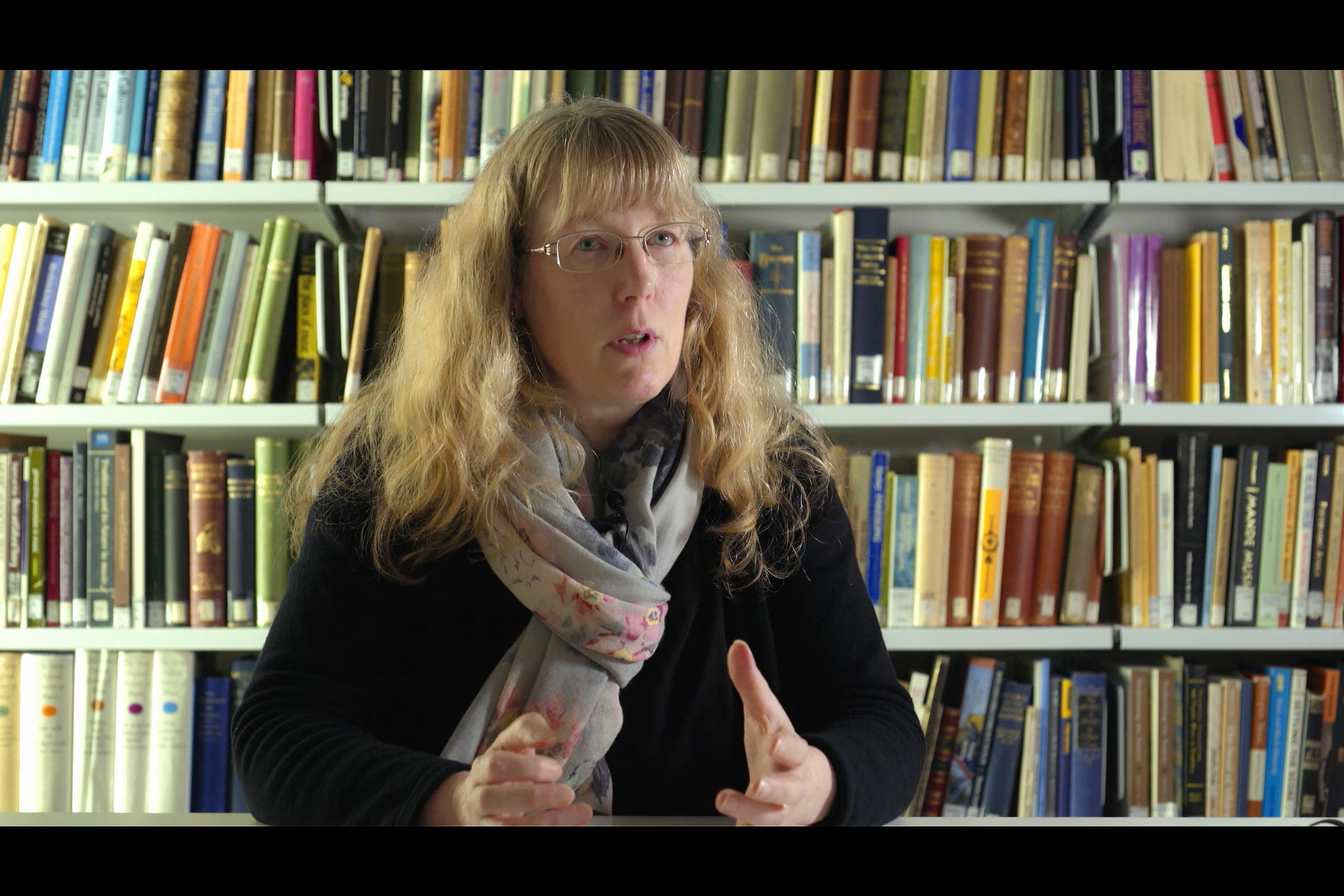 Woman sitting, behind her books on shelves.