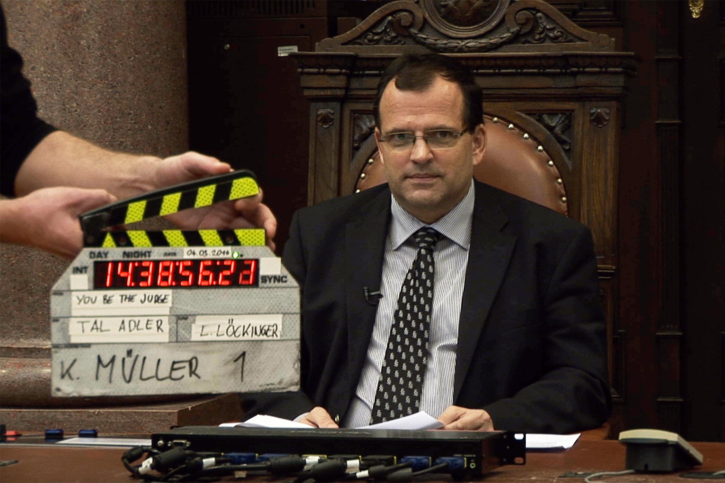 Man sitting on a large decorated wooden chair with leather back. In front of him hands holding a digital movie clapperboard with the date 04.05.2016 and the texts 'YOU BE THE JUDGE', 'TAL ADLER', 'L. LÖCKINGER', and 'K. MÜLLER 1' written on the clapperboard.