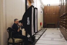 Man sitting next to photography and lighting equipment connected to rails on the floor, in front of a cabinet in a long and narrow hallway