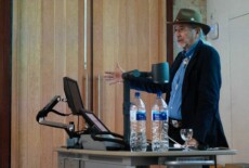 James Riding In speaking at the Dead Images Conference, Edinburgh College of Art, July 2018. Man standing and talking next to a podium in an event hall.