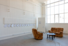 White brochures hanging from hooks in a white wall in an exhibition space with armchairs and a table with printed material on it.