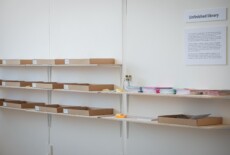 Shelves with boxes and printed material and a sign on the wall with the title 'Unfinished Library'