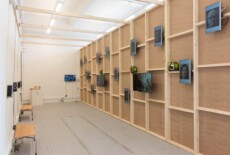 Wooden wall with several video monitors mounted on it and playing different videos at an exhibition space.