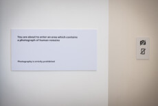 White wall with a sign reading 'You are about to enter an area which contains a photograph of human remains Photography is strictly prohibited.'