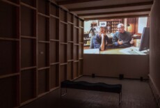 A dark exhibition space with a large video projection on the wall and a bench in front of it.