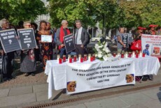 Vigil preceding a repatriation ceremony of Nama and Herero human remains, Berlin, 2018. People standing outside, some holding signs about repatriation. In front of them a table with a banner about the Ovaherero and Nama genocide of 1904-1908.