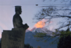 Western Cemetery, Innsbruck, Tyrol, 2012 Out of focus silhouette of a sculpture of a man's bust, Alpine mountains and sunset lit clouds in the background. In the foreground and in focus is a long bubble level.