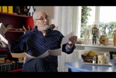Israel Kaunatjike. Still from the video PHOTOGRAPH, part of the Dead Images exhibition, 2018. A man sitting, holding a long collapsible print with both hands, looking at the camera.